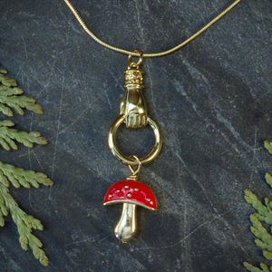 Pode incluir: Um colar de ouro com um pingente de mão e um pingente de cogumelo vermelho. O colar está sobre um fundo cinza escuro.