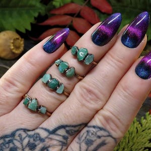 May include: Close-up of several copper-colored rings, each adorned with three raw, green emerald stones. The rings are displayed on a hand with intricate tattoos. The nails are painted with a gradient of blue, purple, and black.