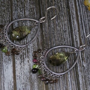 Puede incluir: Un par de pendientes de plata con una piedra preciosa verde en forma de lágrima en cada uno. Los pendientes tienen una cadena delicada con pequeñas piedras preciosas verdes, marrones y rosas.