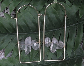 May include: Gold hoop earrings with clear quartz crystals hanging from the hoops. The earrings are on a green leafy background.