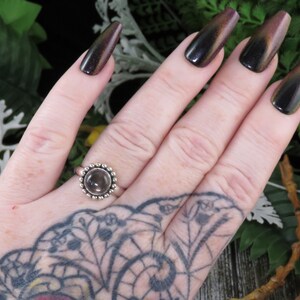 K&ouml;nnte beinhalten: Ein silberner Ring mit einem klaren Edelstein, der in einer Perlenfassung sitzt. Der Ring befindet sich an einer Hand mit dunklem Nagellack.