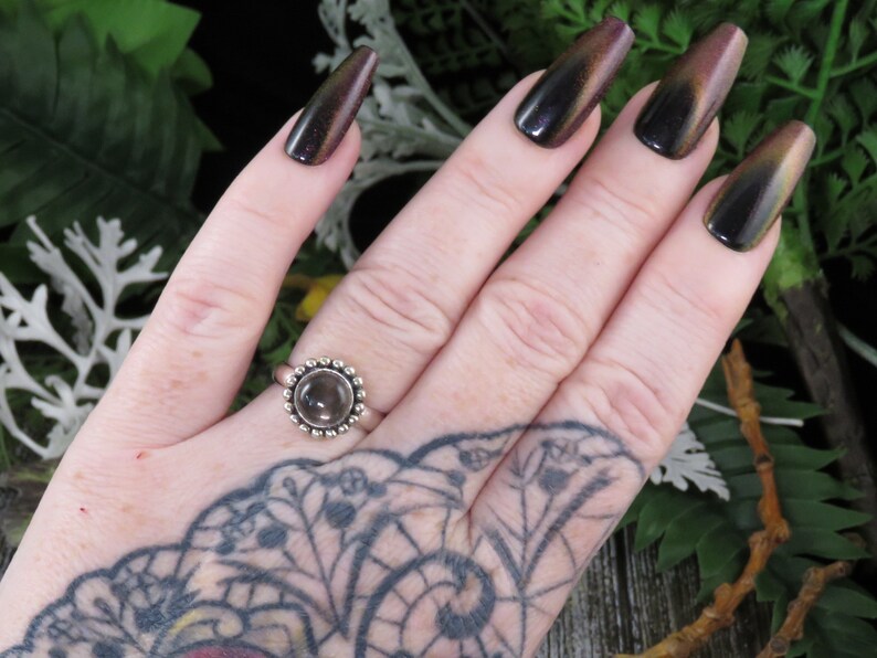K&ouml;nnte beinhalten: Ein silberner Ring mit einem klaren Edelstein, der in einer Perlenfassung sitzt. Der Ring befindet sich an einer Hand mit dunklem Nagellack.