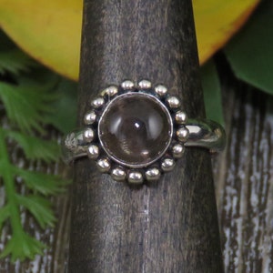 K&ouml;nnte beinhalten: Ein silberner Ring mit einem runden, klaren Edelstein, der in einer Perlenfassung sitzt. Der Ring befindet sich auf einem Holzst&auml;nder.