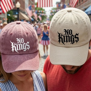 May include: Two baseball caps, one dusty rose and one beige, each with the words "NO KINGS" in a gothic font. The caps are worn by two people in a crowd, with American flags in the background.