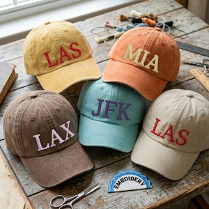 May include: Five baseball caps in various colors, including yellow, orange, brown, light blue, and beige. Each cap features embroidered letters spelling out city abbreviations: LAS, MIA, LAX, JFK, and LAS. The caps are arranged on a wooden surface.