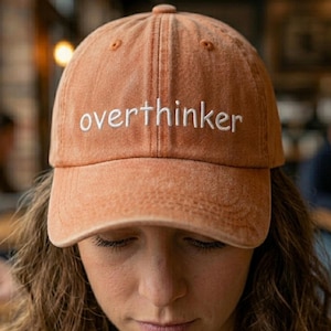 May include: A burnt orange baseball cap with the word "overthinker" embroidered in white. The cap has a curved brim and a classic six-panel design. The hat is made of a soft, textured fabric.