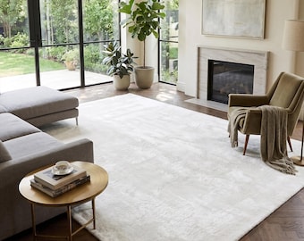 May include: A bright, airy living room with a large, off-white rug. The room features a gray sectional sofa, a small gold side table, and a green armchair. Large windows offer a view of the outdoors, and a fireplace adds warmth.