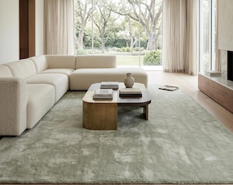 May include: A modern living room with a large, light green rug. A cream-colored sectional sofa sits on the rug, along with a coffee table with a brass base. A large window offers a view of trees and a fireplace.