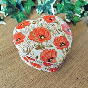 Könnte beinhalten: Herzförmige Schmuckschatulle mit floralem Design. Die Schatulle hat einen weißen Hintergrund mit roten Mohnblumen und kleinen weißen Gänseblümchen. Deckel und Seiten sind mit dem gleichen Muster verziert. Die Schatulle steht auf einer Holzoberfläche.