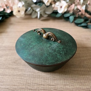 Könnte beinhalten: Ein kleiner, runder, dunkelgrüner Behälter mit einem dekorativen Messinggriff. Der Behälter hat eine strukturierte Oberfläche und einen passenden Deckel. Der Griff ist mit geschwungenen Details verziert. Der Behälter wird wahrscheinlich zur Aufbewahrung oder Präsentation verwendet.