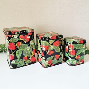 May include: A trio of black metal storage tins adorned with a strawberry design. The tins display red strawberries, green leaves, and white flowers on a black backdrop. Each tin is rectangular and has a lid.