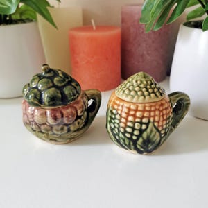 Puede incluir: Dos pequeños recipientes de cerámica con forma de bellota y tapas. Uno es verde y marrón, el otro es verde y naranja. Ambos tienen asas pequeñas y están sobre una superficie blanca. Velas y plantas en el fondo.