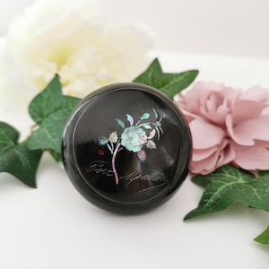 Peut inclure: Boîte décorative ronde noire avec un motif floral aux couleurs irisées. La boîte porte l'inscription "Fine Memories". La boîte est entourée de fleurs blanches et roses et de feuilles vertes.