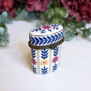 Pode incluir: Caixa de bugigangas oval com tampa articulada. A base branca apresenta desenhos florais e de folhas azuis, com listras douradas. A tampa é decorada com um desenho floral. A dobradiça é de metal cor de latão.