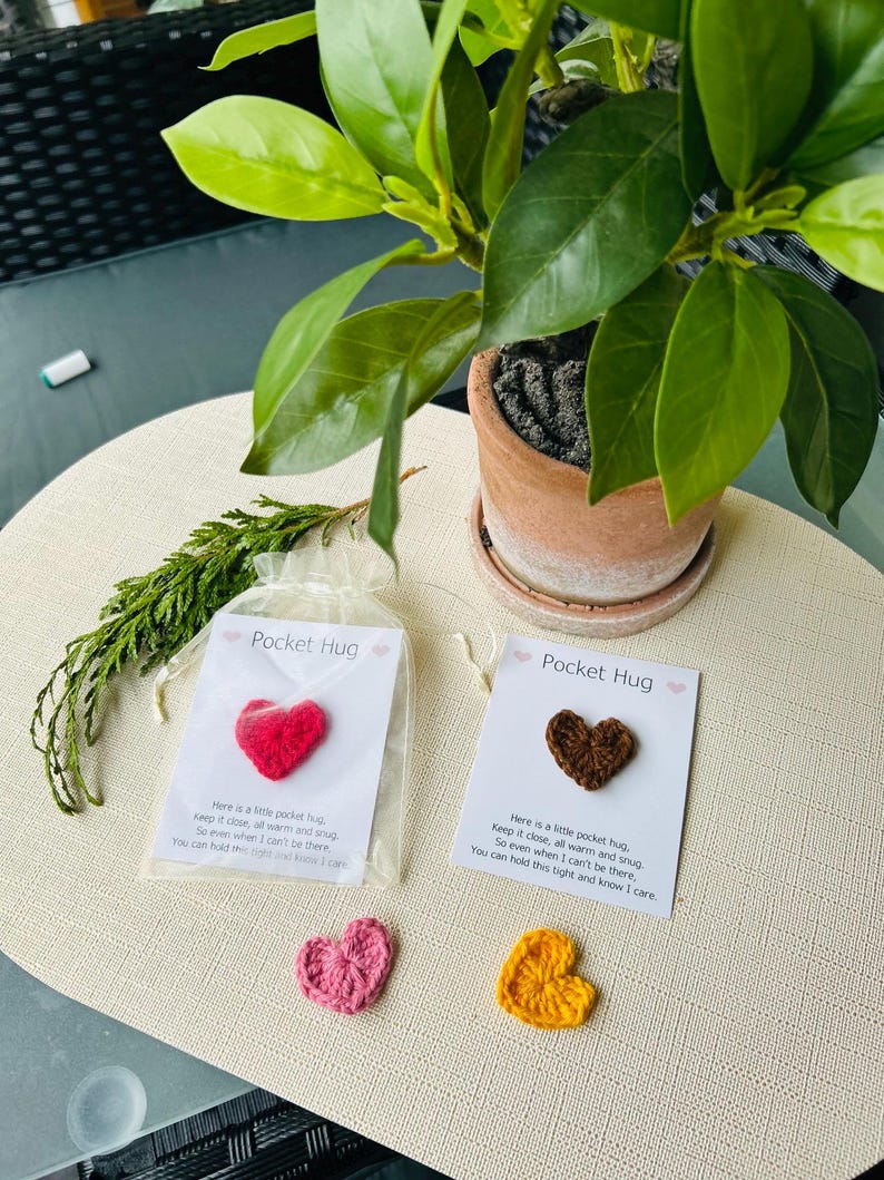 Op de afbeelding: Handgemaakte gehaakte hartvormige Pocket Hugs in rood, bruin, roze en geel. Elk hart wordt gepresenteerd met een kaart met de tekst "Pocket Hug" en een bericht. Een potplant staat op de achtergrond.