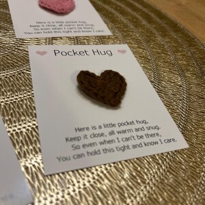 Op de afbeelding: Twee kaarten met de tekst "Pocket Hug" en een klein gehaakt hart. E&eacute;n hart is roze, de andere is bruin. De kaarten bevatten een boodschap over het dichtbij houden van de knuffel. De achtergrond is een goudkleurig, getextureerd oppervlak.