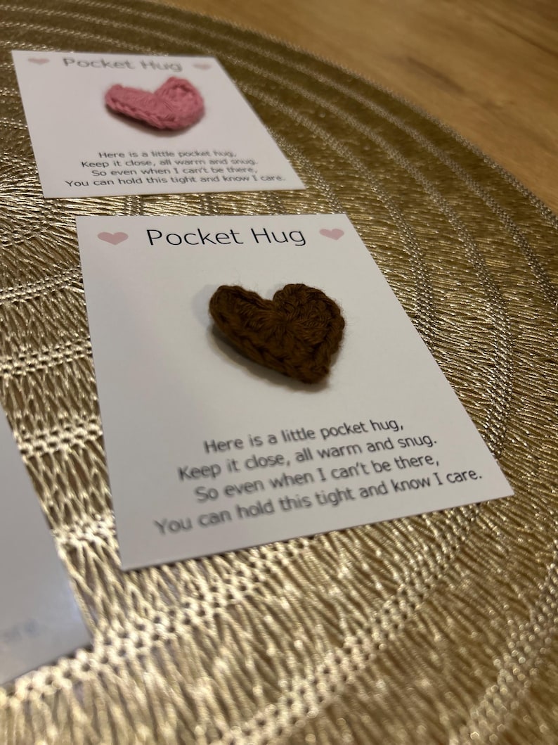 Op de afbeelding: Twee kaarten met de tekst "Pocket Hug" en een klein gehaakt hart. E&eacute;n hart is roze, de andere is bruin. De kaarten bevatten een boodschap over het dichtbij houden van de knuffel. De achtergrond is een goudkleurig, getextureerd oppervlak.
