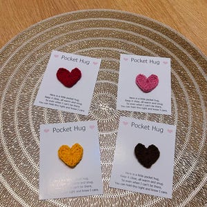 Op de afbeelding: Vier kaarten met gehaakte hartvormige Pocket Hugs in rood, roze, geel en bruin. Elke kaart heeft de tekst "Pocket Hug" en een korte boodschap. De kaarten liggen op een goudkleurige, getextureerde placemat.
