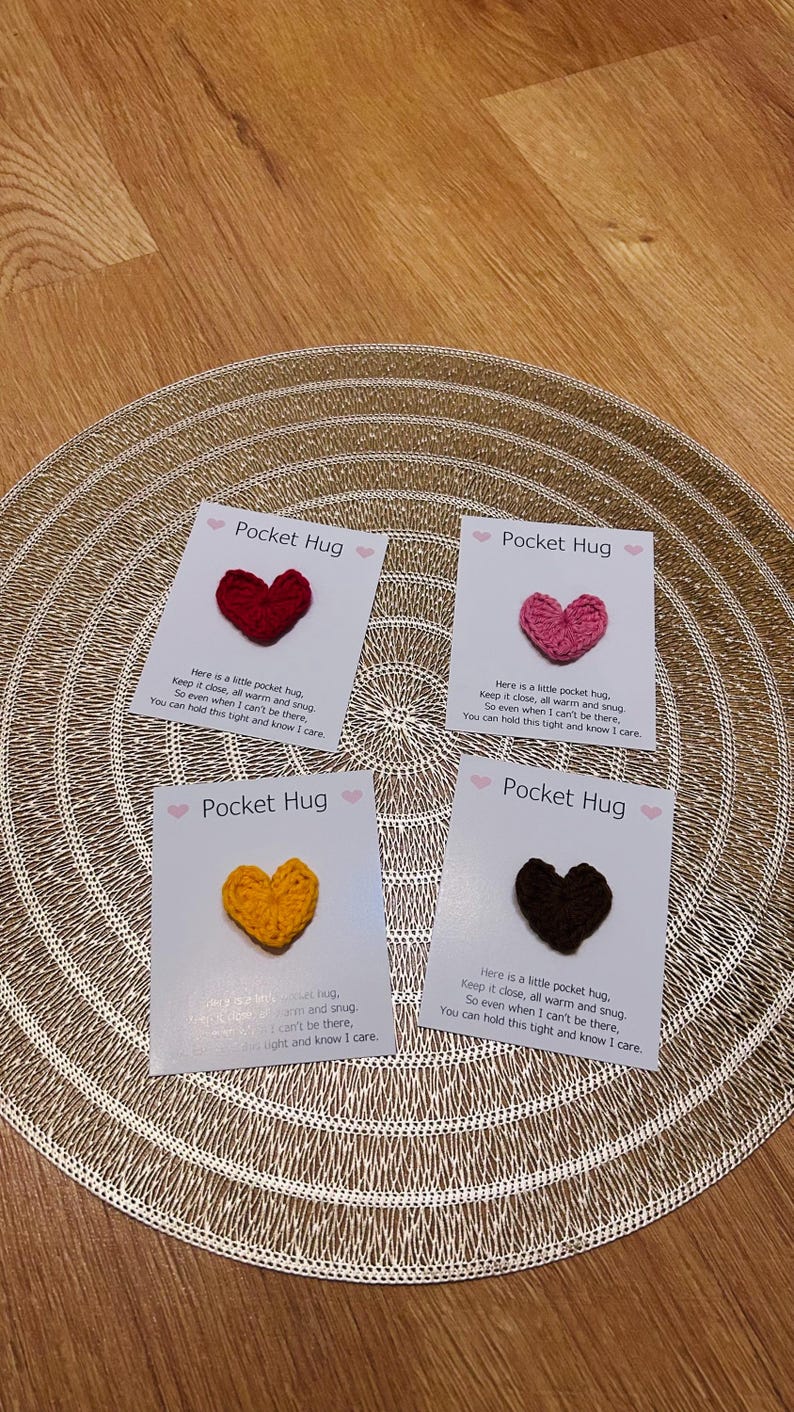 Op de afbeelding: Vier kaarten met gehaakte hartvormige Pocket Hugs in rood, roze, geel en bruin. Elke kaart heeft de tekst "Pocket Hug" en een korte boodschap. De kaarten liggen op een goudkleurige, getextureerde placemat.