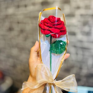 May include: A handcrafted red crochet rose with green leaves, wrapped in clear cellophane and tied with a gold ribbon. The stem is green. A unique, handmade floral gift.