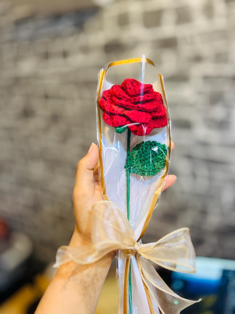 May include: A handcrafted red crochet rose with green leaves, wrapped in clear cellophane and tied with a gold ribbon. The stem is green. A unique, handmade floral gift.