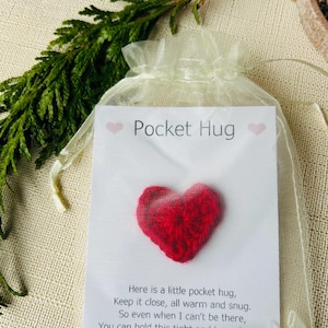 Op de afbeelding: Een klein, rood, hartvormig item in een doorzichtige organza zak. De zak bevat een kaart met de woorden "Pocket Hug" en een gedicht. De achtergrond bevat groen en een neutraal gekleurd oppervlak.