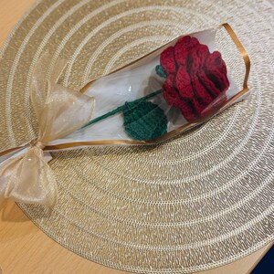 May include: A handcrafted red crochet rose with a green leaf and stem, presented in a clear cellophane wrapper. The stem is tied with a gold ribbon. The rose is displayed on a gold-colored placemat.