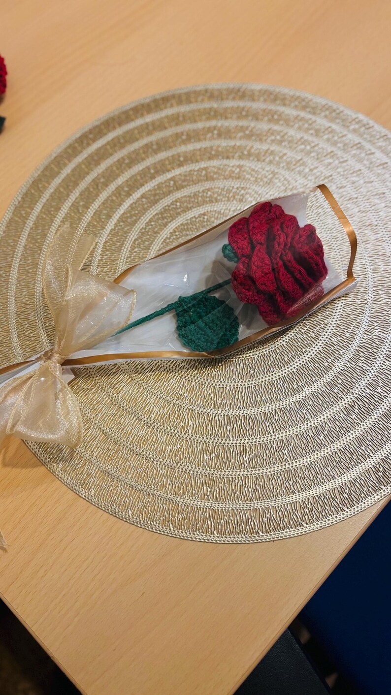 May include: A handcrafted red crochet rose with a green leaf and stem, presented in a clear cellophane wrapper. The stem is tied with a gold ribbon. The rose is displayed on a gold-colored placemat.