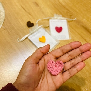 Op de afbeelding: Een roze gehaakt hart wordt in een hand gehouden, met twee andere hartvormige hangers in kleine zakjes op de achtergrond. De zakjes bevatten een gele en een rode hartvormige hanger. De achtergrond is een houten oppervlak.