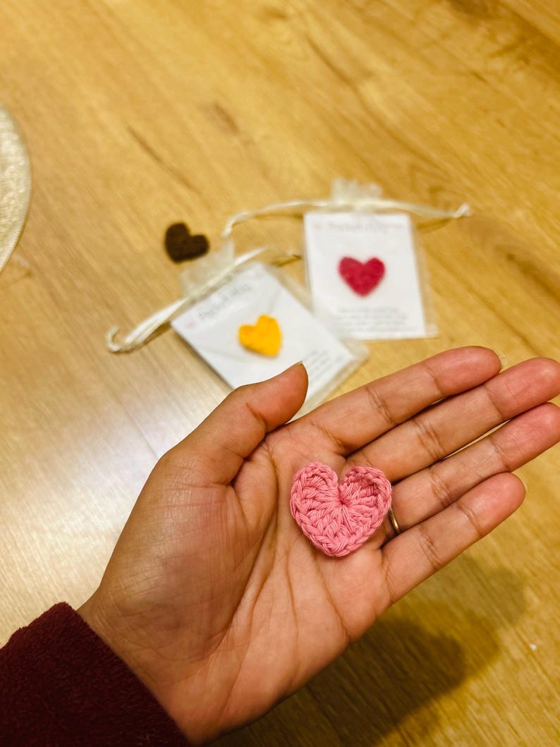 Op de afbeelding: Een roze gehaakt hart wordt in een hand gehouden, met twee andere hartvormige hangers in kleine zakjes op de achtergrond. De zakjes bevatten een gele en een rode hartvormige hanger. De achtergrond is een houten oppervlak.
