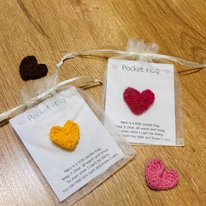 Op de afbeelding: Verschillende gehaakte hartvormige items worden getoond. Twee zitten in kleine, transparante zakjes met de tekst "Pocket Hug" en een bericht. Extra harten in bruin en roze zijn ook zichtbaar. De harten liggen op een houten oppervlak.