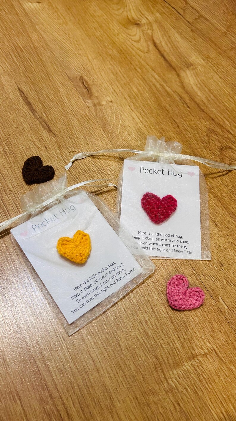 Op de afbeelding: Verschillende gehaakte hartvormige items worden getoond. Twee zitten in kleine, transparante zakjes met de tekst "Pocket Hug" en een bericht. Extra harten in bruin en roze zijn ook zichtbaar. De harten liggen op een houten oppervlak.