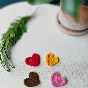 Op de afbeelding: Vier kleine, gehaakte harten in rood, geel, bruin en roze zijn gerangschikt op een wit, getextureerd oppervlak. Een takje groen loof en een terracotta pot met een plant staan op de achtergrond. De harten zijn waarschijnlijk handgemaakt.