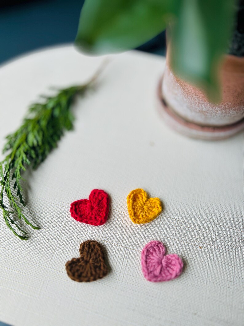 Op de afbeelding: Vier kleine, gehaakte harten in rood, geel, bruin en roze zijn gerangschikt op een wit, getextureerd oppervlak. Een takje groen loof en een terracotta pot met een plant staan op de achtergrond. De harten zijn waarschijnlijk handgemaakt.