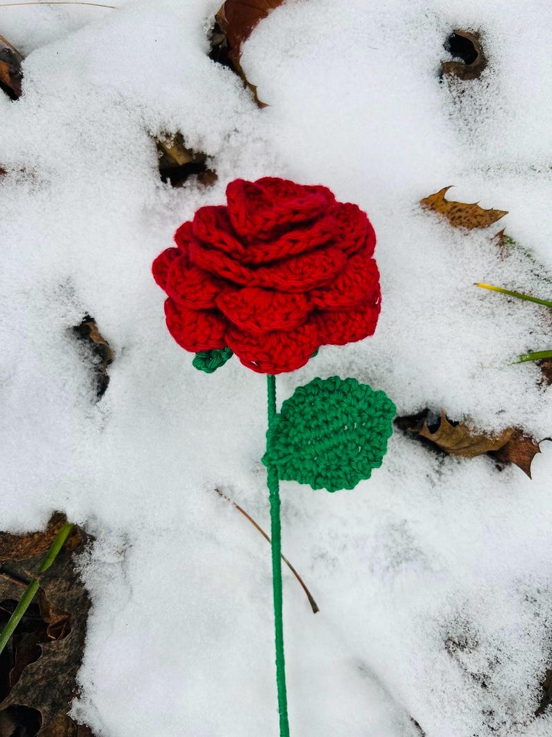May include: A handmade crochet rose with a vibrant red bloom, green stem, and leaf. The flower is set against a snowy backdrop, showcasing the intricate details of the yarn work. A unique, handcrafted floral accent.