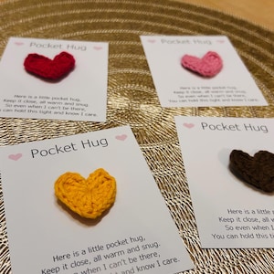 Op de afbeelding: Vier kaarten met gehaakte hartvormige Pocket Hugs in rood, roze, geel en bruin. Elke kaart heeft de woorden "Pocket Hug" en een korte boodschap. De kaarten liggen op een gouden, getextureerd oppervlak.