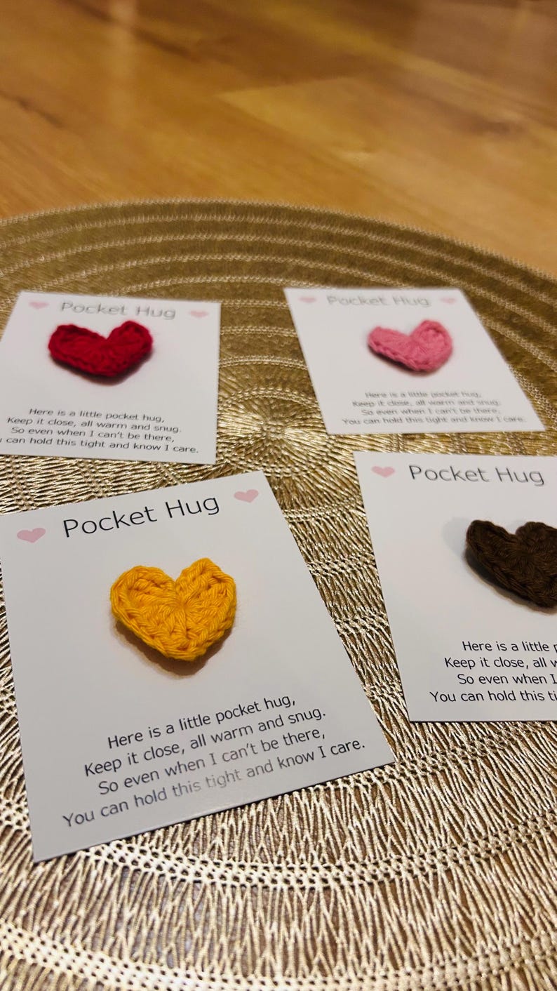 Op de afbeelding: Vier kaarten met gehaakte hartvormige Pocket Hugs in rood, roze, geel en bruin. Elke kaart heeft de woorden "Pocket Hug" en een korte boodschap. De kaarten liggen op een gouden, getextureerd oppervlak.