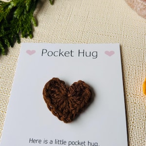 Op de afbeelding: Een bruin gehaakt hart op een witte kaart met de tekst "Pocket Hug". De kaart bevat ook een gedicht over een zakknuffel. De achtergrond omvat groen en een beige oppervlak.
