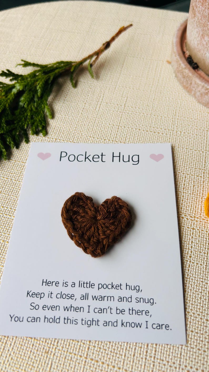 Op de afbeelding: Een bruin gehaakt hart op een witte kaart met de tekst "Pocket Hug". De kaart bevat ook een gedicht over een zakknuffel. De achtergrond omvat groen en een beige oppervlak.