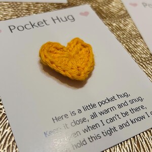 Op de afbeelding: Een handgemaakt, geel gehaakt hart op een witte kaart met de tekst "Pocket Hug". De kaart bevat ook een bericht. Andere kaarten met rode en roze harten zijn zichtbaar.