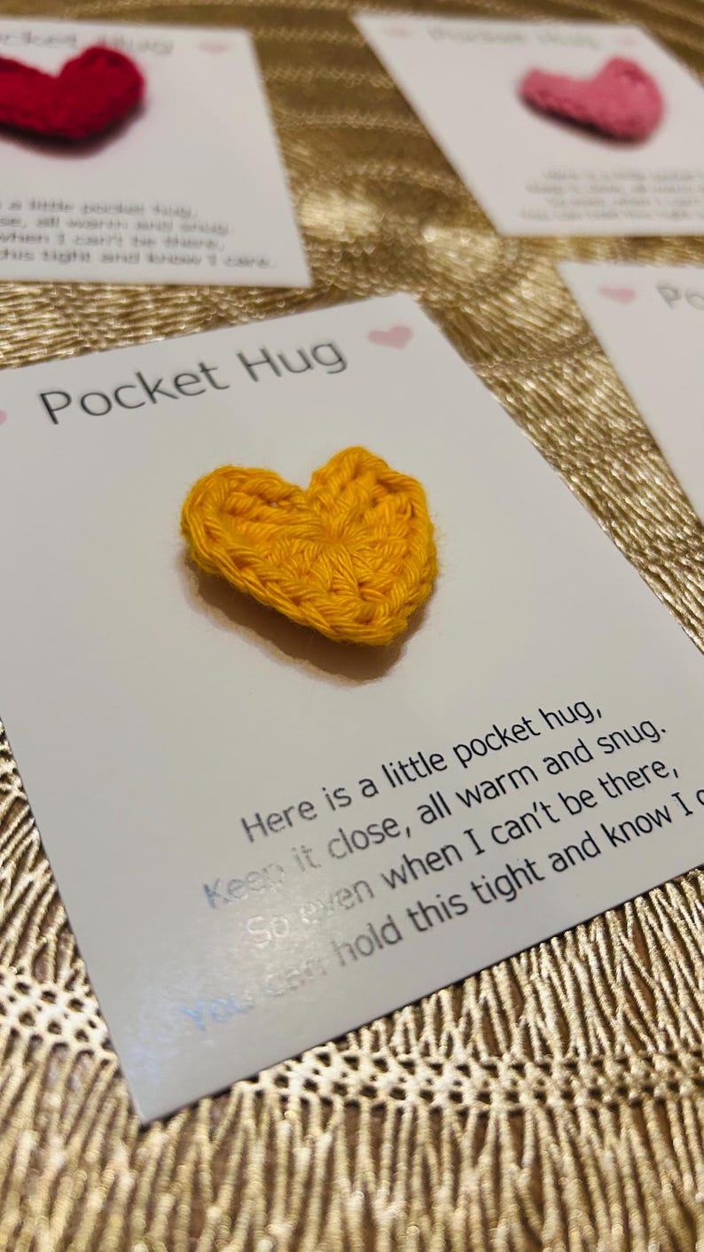 Op de afbeelding: Een handgemaakt, geel gehaakt hart op een witte kaart met de tekst "Pocket Hug". De kaart bevat ook een bericht. Andere kaarten met rode en roze harten zijn zichtbaar.