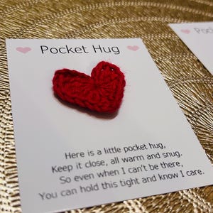 Op de afbeelding: Een rood gehaakt hart ligt op een witte kaart met de woorden "Pocket Hug" en een bericht. De kaart ligt op een goudkleurig, getextureerd oppervlak. Het hart is een klein, handgemaakt item.