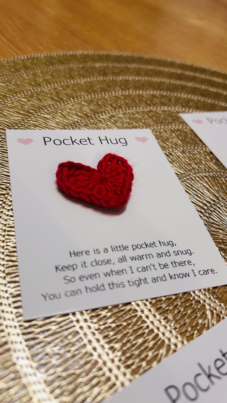 Op de afbeelding: Een rood gehaakt hart ligt op een witte kaart met de woorden "Pocket Hug" en een bericht. De kaart ligt op een goudkleurig, getextureerd oppervlak. Het hart is een klein, handgemaakt item.