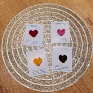 Op de afbeelding: Vier kleine kaarten met gehaakte hartvormige Pocket Hugs in rood, roze, geel en bruin. Elke kaart heeft de tekst "Pocket Hug" en een korte boodschap. De kaarten zijn gerangschikt op een goudkleurige placemat.