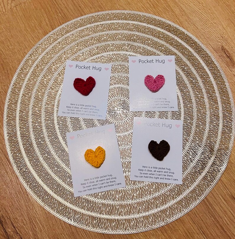 Op de afbeelding: Vier kleine kaarten met gehaakte hartvormige Pocket Hugs in rood, roze, geel en bruin. Elke kaart heeft de tekst "Pocket Hug" en een korte boodschap. De kaarten zijn gerangschikt op een goudkleurige placemat.