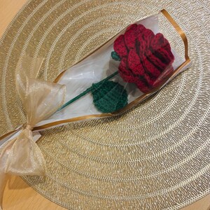 May include: A handcrafted red crochet rose with a green stem and leaf, wrapped in clear cellophane and tied with a gold ribbon. The rose is presented on a gold textured placemat, creating a romantic and decorative gift.
