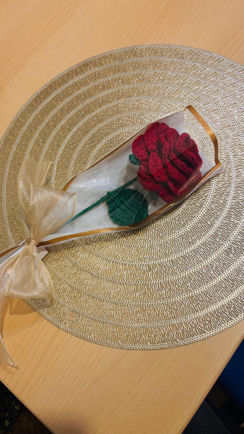 May include: A handcrafted red crochet rose with a green stem and leaf, wrapped in clear cellophane and tied with a gold ribbon. The rose is presented on a gold textured placemat, creating a romantic and decorative gift.