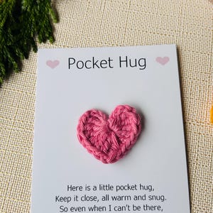 Op de afbeelding: Een roze gehaakt hart op een witte kaart met de tekst "Pocket Hug". De kaart bevat ook een gedicht over een zakknuffel. De achtergrond bevat een takje groen.