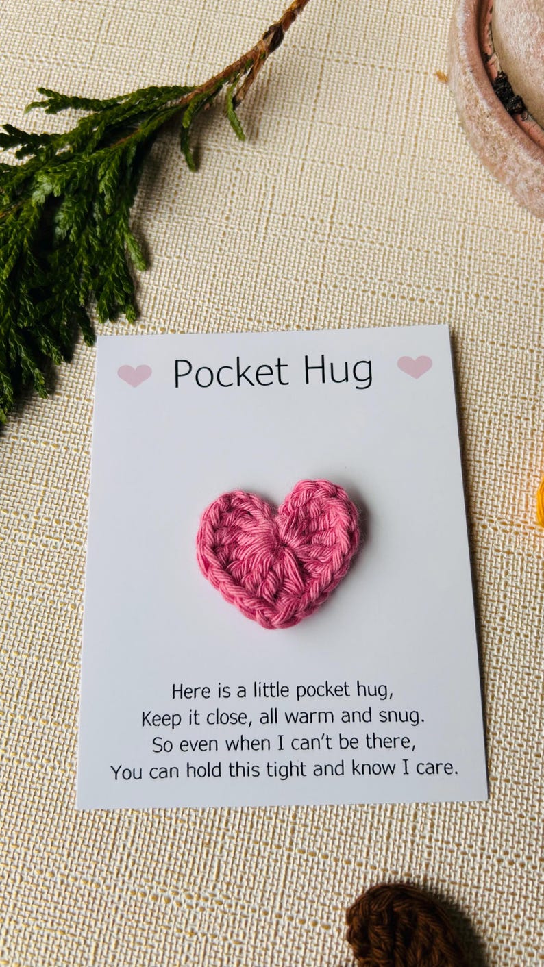 Op de afbeelding: Een roze gehaakt hart op een witte kaart met de tekst "Pocket Hug". De kaart bevat ook een gedicht over een zakknuffel. De achtergrond bevat een takje groen.