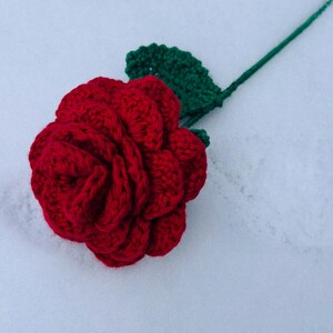 May include: A handmade, red crochet rose with a green leaf and stem. The flower is in full bloom, with detailed petals. The stem and leaf are a vibrant green. The rose is a craft item, possibly for decoration or as a gift.